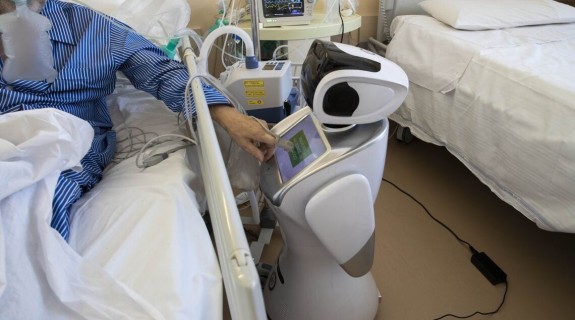 A patient in a hospital bed reaches out to press buttons on a hospital robot