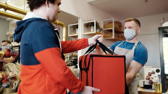 A person wearing a mask and an apron hands an orange food delivery bag to a person wearing a blue and orange jacket