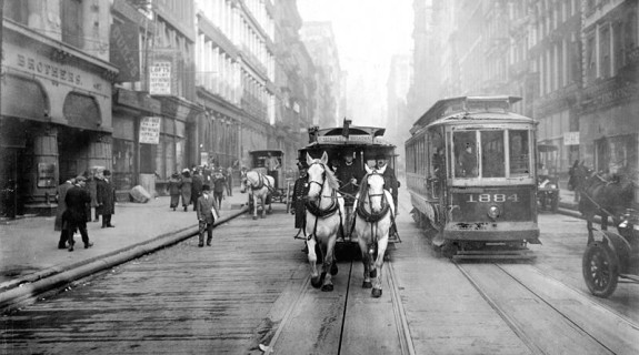Two horses pull a cart on a street next to a trolley with the front label 1884