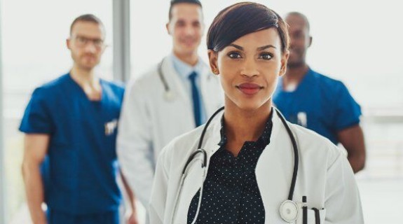 A medium toned woman wearing a white coat and a stethoscope stands in front of 3 other healthcare workers
