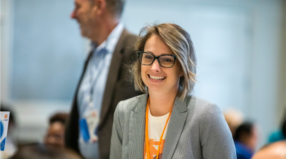 Woman smiling wearing black framed glasses, a gray suit, and a conference lanyard..