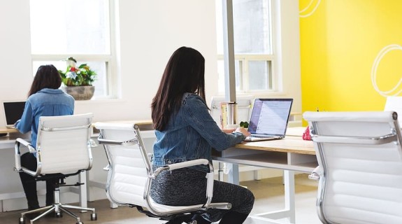 A person in a jean jacket and black and white leggings works at a laptop on a table