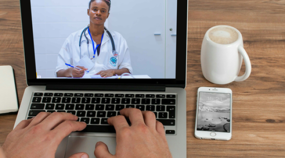 A dark toned woman wearing a white coat and blue stethoscope appears on a laptop screen while two hands type on the keyboard