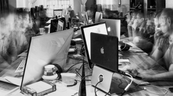 A group of open laptops and computer accessories on two tables surrounded by ghostly people