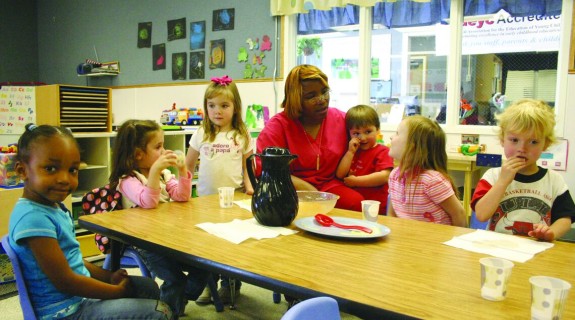 An adult sits at a long wooden table surrounded by children