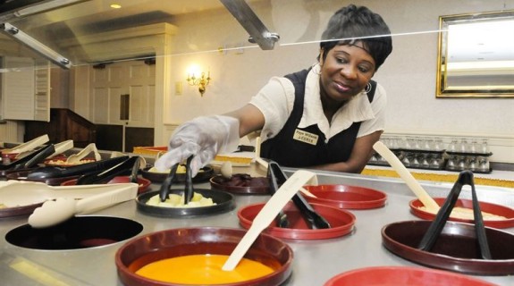 A Black woman reaches across containers of food under a buffet shield