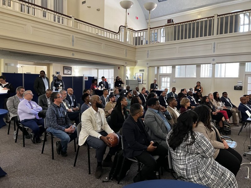 An audience of several dozen people sit in a room listening to a conference presentation