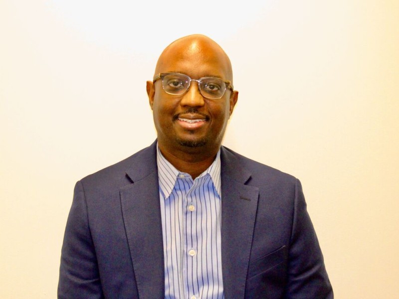 man smiling in blue shirt and navy blazer wearing glasses