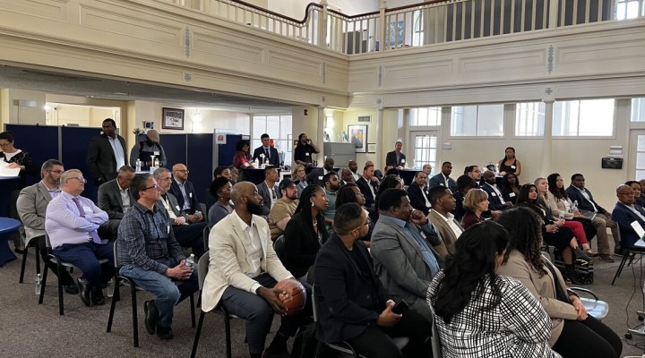 An audience of several dozen people sit in a room listening to a conference presentation